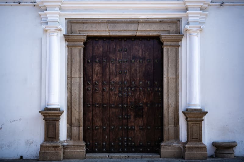 A dark studded wooden double door in a carved stone frame between white columns, in the Spanish colonial style